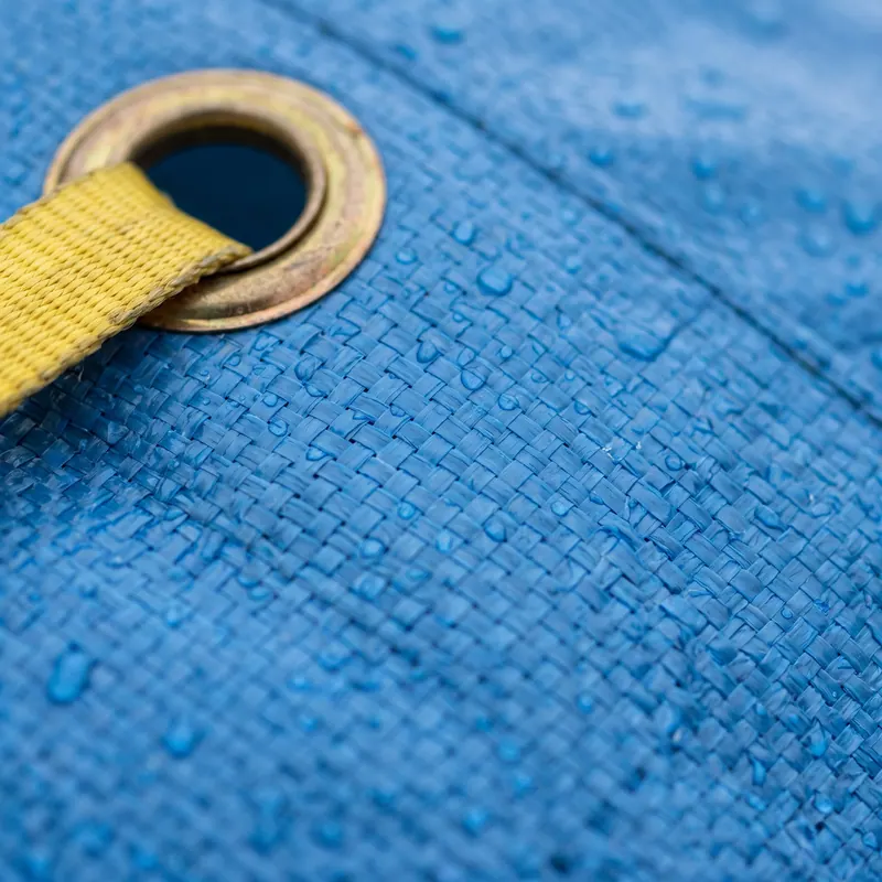 Close-up of a wet blue tarp: rain beaded across the woven polyethylene and a yellow strap threaded through a brass grommet